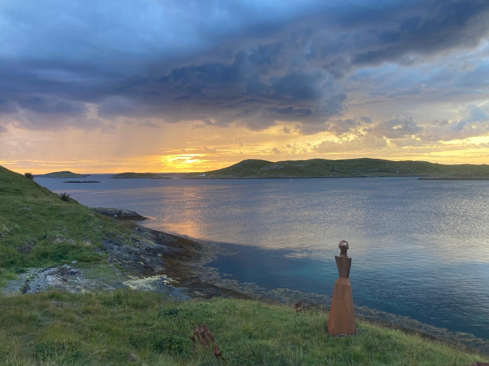 A rust-red sculpture of a woman on the shore, looking out over the sea at sunset