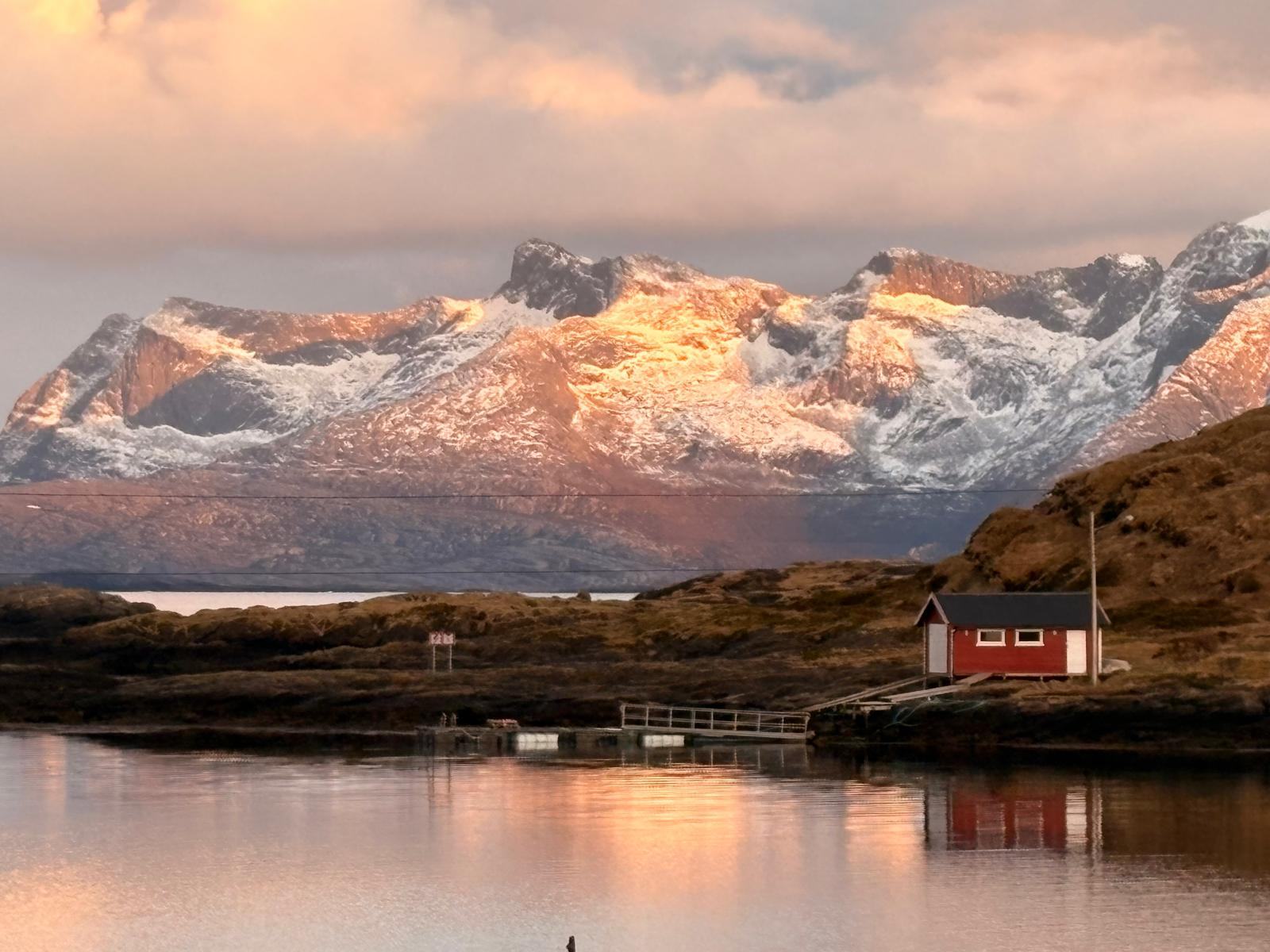 A small red boathouse below snow-covered mountains at dusk